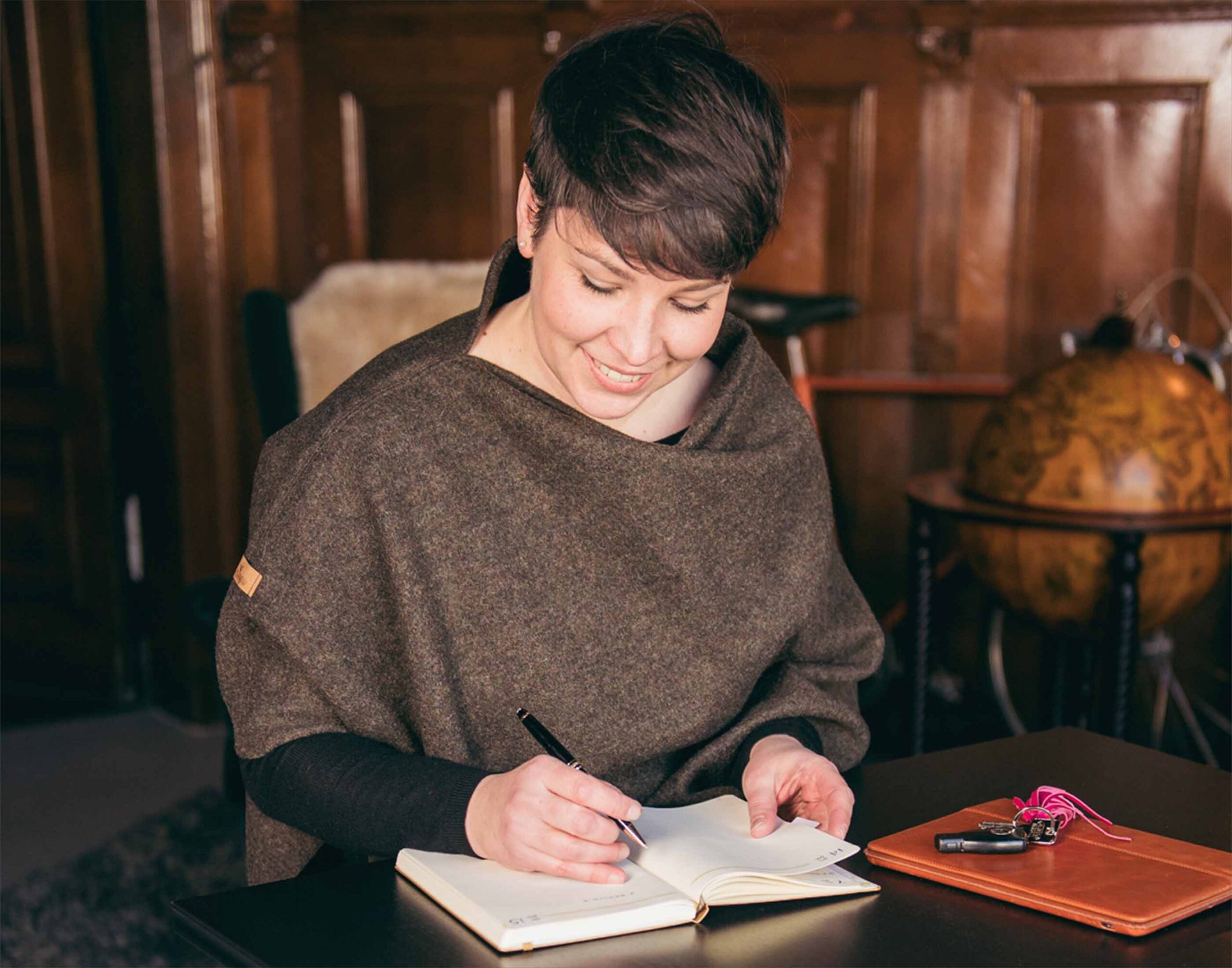 Maren Gutzmann - Autorin Die Autorin sitzt lächelnd an einem Tisch und schreibt in ein Notizbuch. Sie hat kurze, dunkle Haare und trägt einen dunkelbraunen Poncho.
