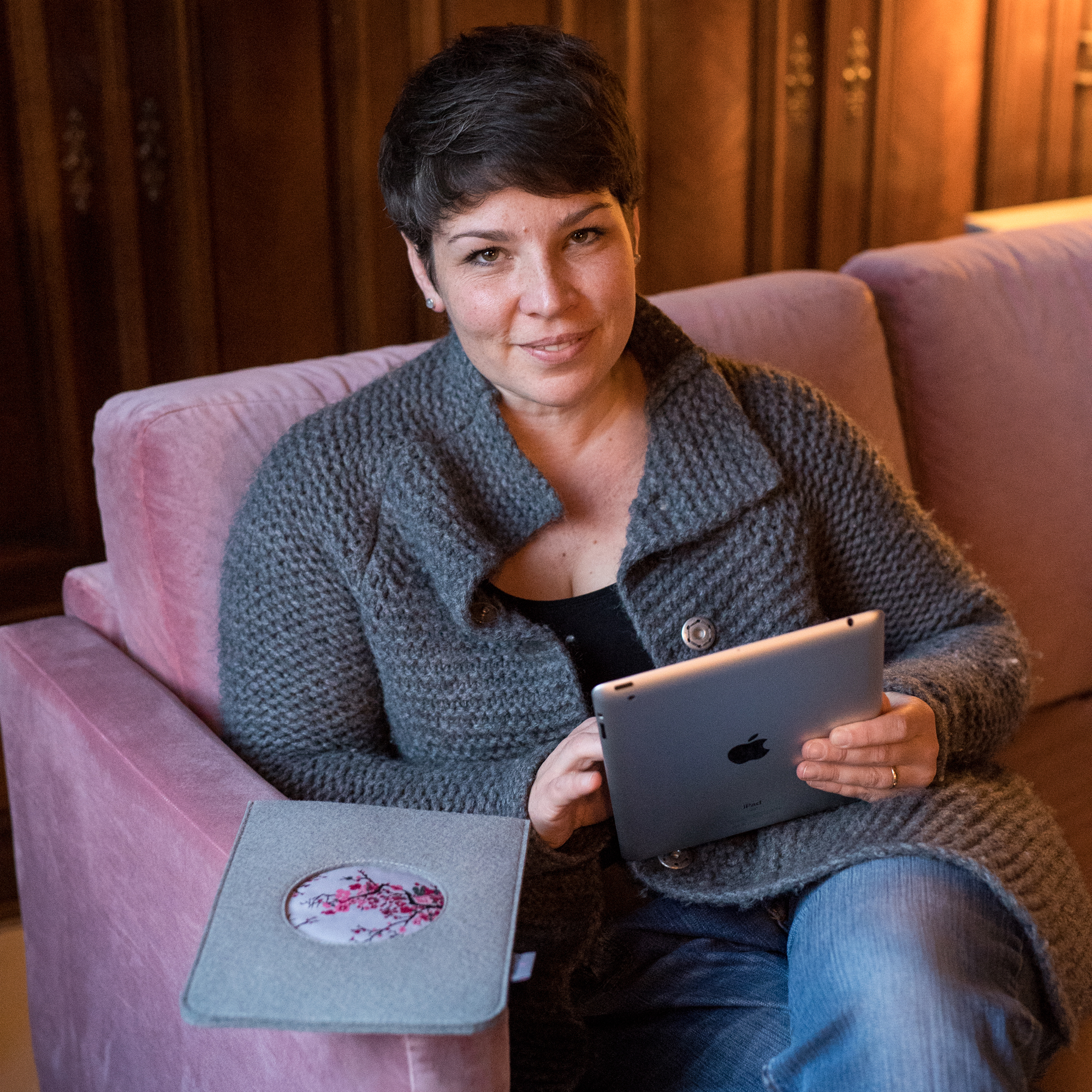 Autorin Maren Gutzmann Die Autorin sitzt auf einem rosafarbenen Sofa mit einem Tablet in der Hand. Sie hat kurze braune Haare, lächelt in die Kamera und trägt eine dunkelgraue Strickjacke.